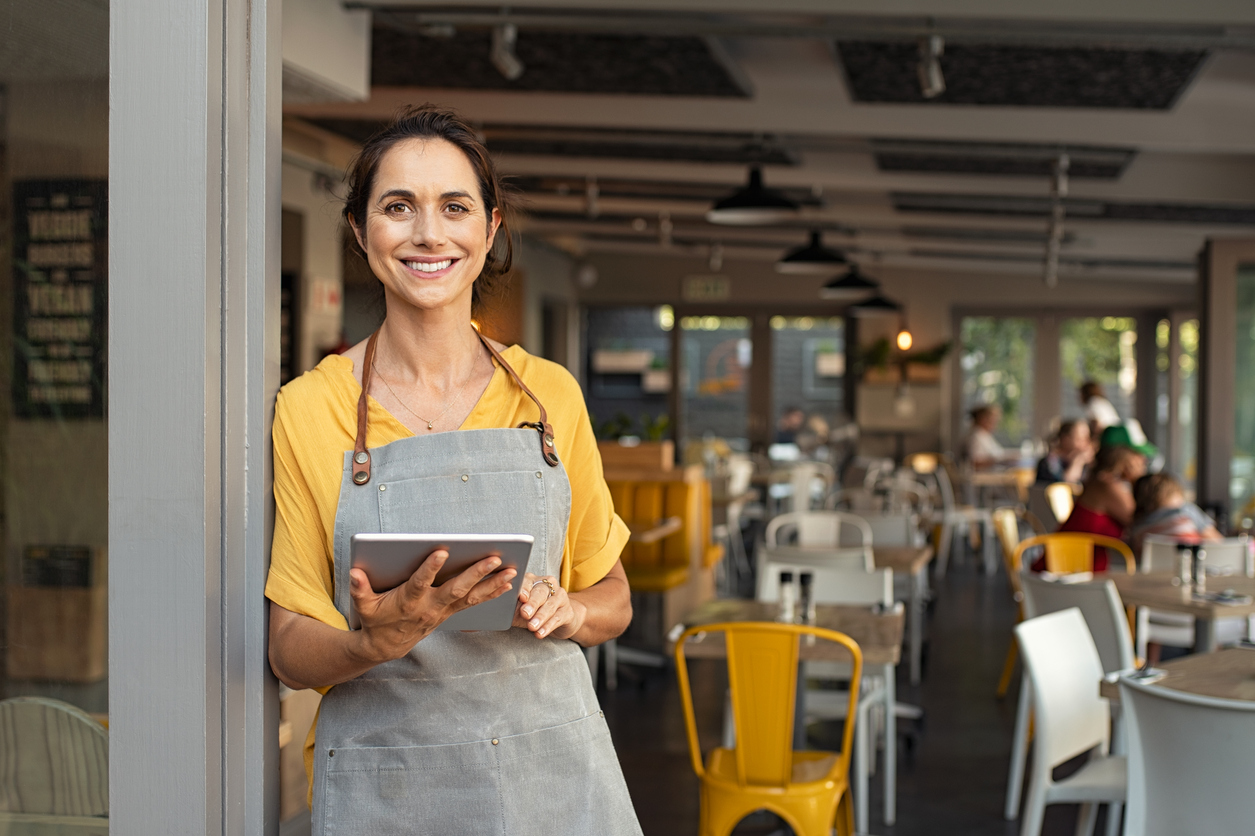 Female restaurant owner standing at the doorway while holding a tablet device to manage her NY restaurant payroll and other employee needs.